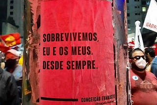 Lambe vermelho com a frase “sobrevivemos, eu e os meus. Desde sempre.” da Conceição Evaristo, colada num poste. Ao redor as pessoas erguem os punhos em meio a um protesto.