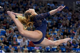 Image Description: Female gymnast Katelyn Ohashi wearing a navy blue leotard doing a split in the air during a balance beam routine. Photo credit: UCLA Gymnastics. https://uclabruins.com/sports/womens-gymnastics