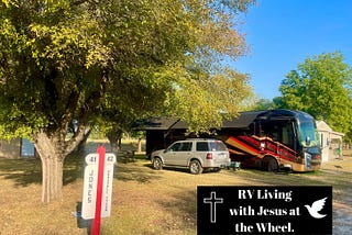 Big Red, our black and red Entegra Anthem motorhome, parked beneath shady trees at our RVICS Village home base in Smithville, Texas, with our site marker and RV Living with Jesus at the Wheel logo in the foreground on a peaceful sunny afternoon.