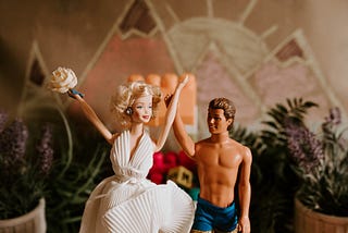 A Barbie doll wearing a white dress and holding a bouquet. She is standing beside a Ken doll wearing only board shorts.