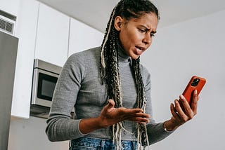 A woman holds a red smartphone and appears frustrated or angry at it.