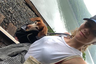 Rebecca, a white woman with curly hair and a Mets hat on, smiles while she carries her brown dachshund, Forrest, in a bag.