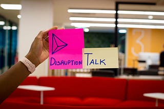 A hand holding up two Post-it notes. On one is written “Disruption” with a doodle of a pyramid. On the other is written “Talk.”