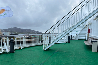 Deck of a Wales to Ireland ferry, with green astro turf, white railings and misty mountains in the distance. A flag whips in the wind on a stormy day.