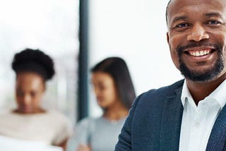 Image of a man in the foreground smiling with 2 other coworkers in the background looking at a document