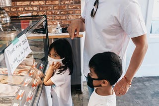 A boy in a mask pointing at a gelato flavor as they look at a wide display of all sorts of flavors