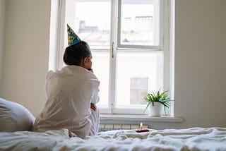 woman sits with back to the camera, wearing a party hat, cupcake with candle next to her