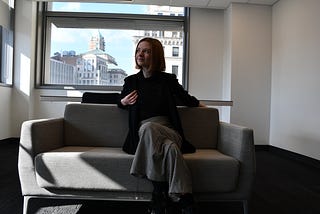 A blonde white woman sitting on a grey couch in front of a window.