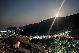 View of moon over distant hill with lights of the village in the valley twinkling int he night and the sea in the distance.