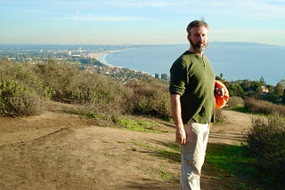 A young bearded man stands on a bluff overlooking a coastline
