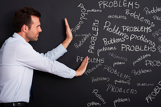 A photo of a man by a blackboard covered with many instances of the word “problems.”