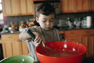 A toddler holding a whisk mixing in a large red bowl.