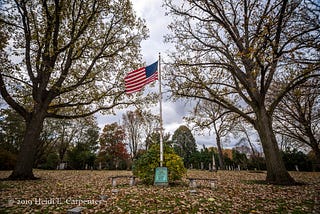 A flagpole with an American flag, flanked by trees, in a cemetery on an overcast autumn day.