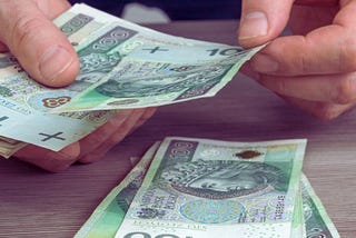 Na zdjęciu widoczne są ręce osoby liczącej polskie banknoty o nominale 100 złotych. W tle po prawej stronie leży kalkulator, co może sugerować, że osoba dokonuje obliczeń związanych z pieniędzmi. Kilka banknotów leży na blacie stołu, a reszta jest trzymana w rękach.