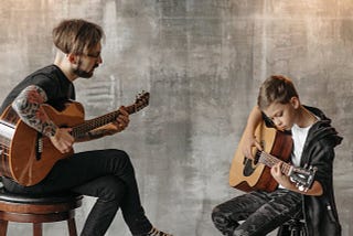 Two men sitting on stools, playing a classical guitar. The older one is teaching the younger one, a pre-teen, how to play guitar.