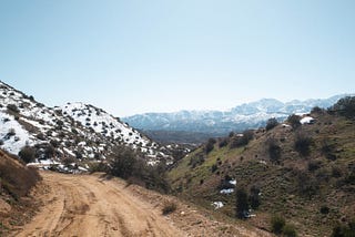 view of a dirt trail winding up a mountain. Looking down the trail and off to the mountains in the distance with snow-capped peaks