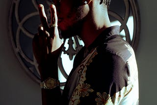 A Black man wearing a stylish shirt and jewelry, confidently holding up a peace sign with his fingers.