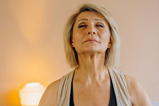 A middle aged woman with shoulder length blond hair wearing a gray tank top. She is breathing with her eyes closed and chin tilted upward facing the camera.