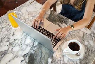 A lady with long light colored hair is sitting at a marble top table. You can only see her body and arms, and she is wearing a sleeveless, dark outfit. Her arms are stretched onto the table. She is working on a silver laptop. To her left is a cup of coffee on a saucer (with a spoon also laying in the saucer), and on her right is a glass of orange juice with a white bendy straw.