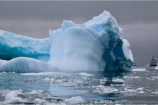 Large bue iceberg with boat nearby in icy waters against gray sky