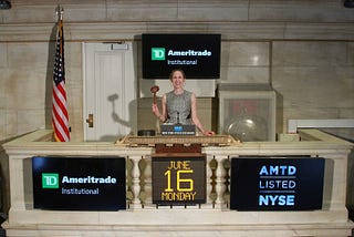 A white woman in a grey dress standing with a gavel, prentending to ring the closing bell at the New York Stock Exchange (NYSE). Monitors feature the TD Ameritrade Institutional logo.