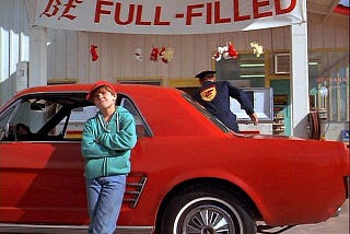 The main character of “Motorama”, Gus, leans against his Mustang convertible at a gas station. The banner in the background reads “Be Full-Filled.”
