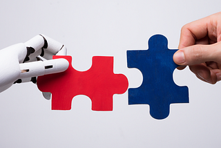 On the left is a robotic hand holding a red puzzle piece. On the right, a human hand holds a blue puzzle piece. The two hands are attempting to fit the puzzle pieces together.