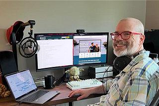 White man with a beard and red glasses smiles as he sits in front of a desk holding a laptop and two monitors.