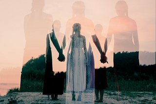 3 women standing on a rock, possibly next to a river, holding hands, facing away from the camera toward a set of trees, silhouette