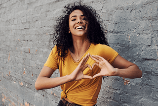 A young woman, standing against a grey backdrop, with her fingers joined together to create a heart shape. It depicts self love.