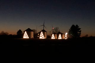 Four grain bins. Three with Christmas lights arranged in the shape of Christmas trees and one in the shape of a snowman.