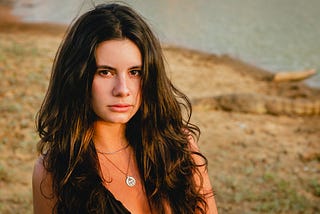 Woman with long dark hair staring at camera in a dark lowcut shirt and silver necklace, in front of a sandy background.