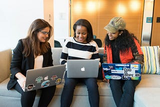 Three young women, possibly college students, sitting on a couch, smiling and conversing, with laptops on their laps.