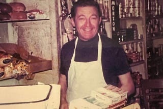 Man wearing white apron at his grocery store, standing near scale