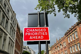 A red road sign with white text reads “CHANGED PRIORITIES AHEAD.” It is mounted on two black poles and stands in an urban street lined with historic brick and stone buildings. A tree with green leaves partially covers the top right corner of the image, while the sky is cloudy. The street appears quiet, with a few pedestrians and a car in the background.