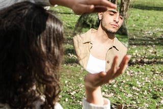 Picture of a couple, with woman holding a mirror and the man’s reflection looking back at her, with grassy knoll in backdrop
