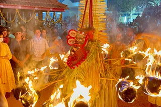 A performer surrounded by flaming torches during a nighttime ritual at a temple, with devotees and attendants watching him in awe.