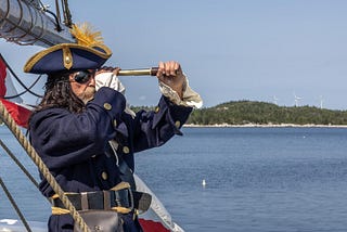 Man in a pirate costume with eye patch and buccaneer hat, aboard a sailing ship, peering through a spyglass, with an island in the background.