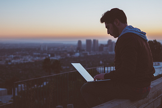 A man on his laptop, sitting on a wall atop a tall building. The backdrop is a city against an orange, darkening sky as the sun sets.