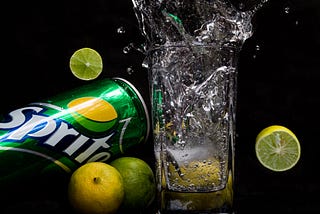 Splash photo of sprite on a black background with lemons in the air and sprite can tilting along with the glass.