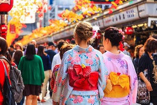 Two women wearing colorful kimono walk along Nakamise Dori near Senso-ji in Tokyo, surrounded by a busy crowd of tourists.