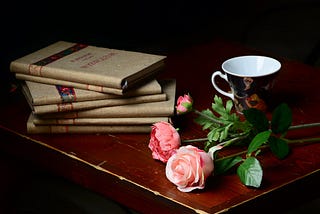 Poetry books set on top of one another next to a tea cup and two pink roses on a wooden table.