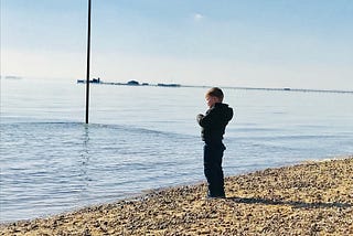 Alfie is standing at the edge of the sea on stony sand looking at something he is holding in his hand. You an see a pole sticking out of the blue sea with ripples around it, and a pier in the distance.