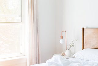 A light-filled bedroom with a bed with white sheets and a side table with a copper accent light and a plant.