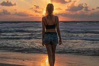 A woman in a black crop top and jean shorts watches the Sun set over the ocean on a beach.