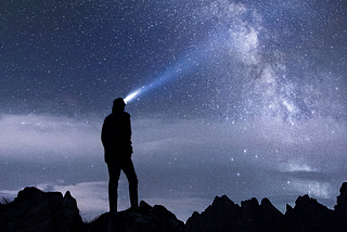 The silhouette of a man and the rocks he’s standing on, with a headlamp on, looking up into the night’s sky. The sky is full of stars with the Milky Way visible across it.