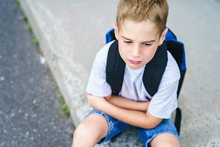 A little boy wearing a backpack sits on the sidewalk looking sad. His arms are loosely crossed in his lap.