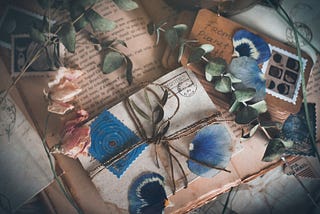 A flat lay of vintage-style correspondence against a soft, moody background. In the center, a small parcel wrapped in aged paper and tied with gold string is adorned with pressed blue flower petals and dried eucalyptus leaves. The parcel rests on a bed of old letters with cursive script, scattered with dried pink roses and decorative postage stamps. The overall mood is nostalgic and gentle.