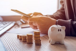 A white piggy bank and stacks of quarters are in the foreground, a person is on their phone in the background.