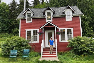 A 70 year old woman on the steps of her farmhouse-style red cottage near the woods.